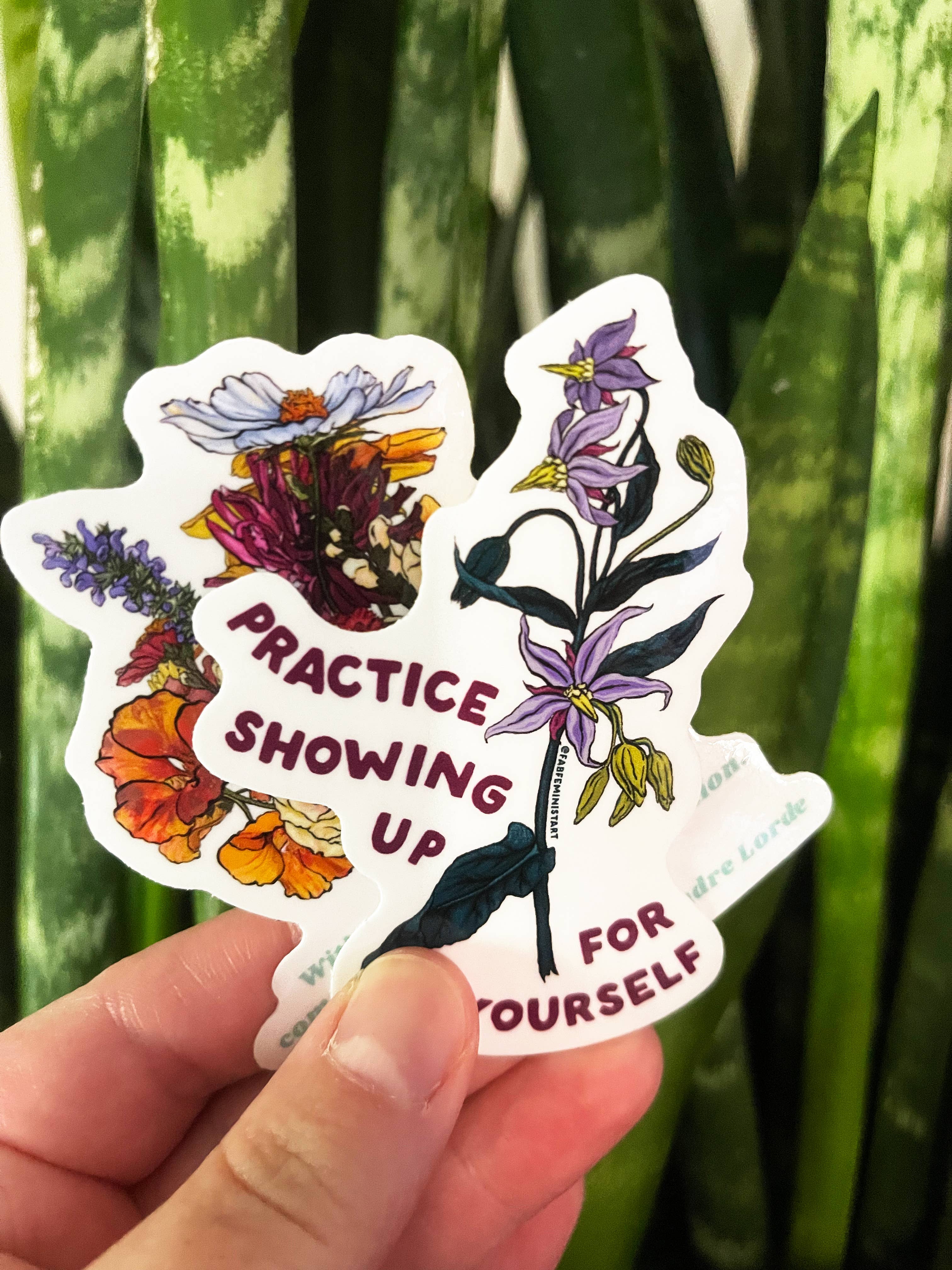 A sticker with a floral design and the text 'PRACTICE SHOWING UP FOR YOURSELF' held against a backdrop of green leaves.