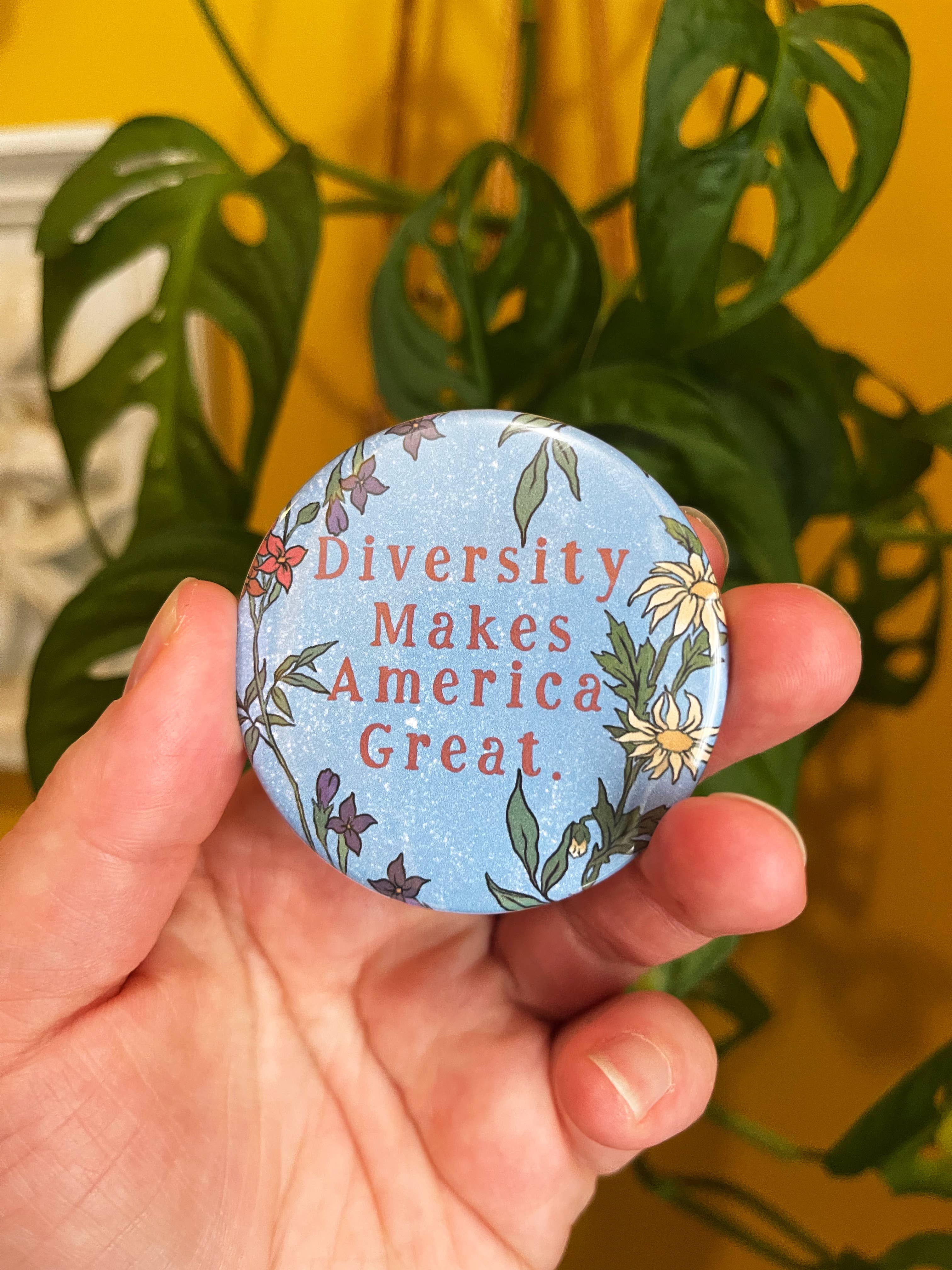 A hand holding a round pinback button with the text 'Diversity Makes America Great' surrounded by a floral pattern.