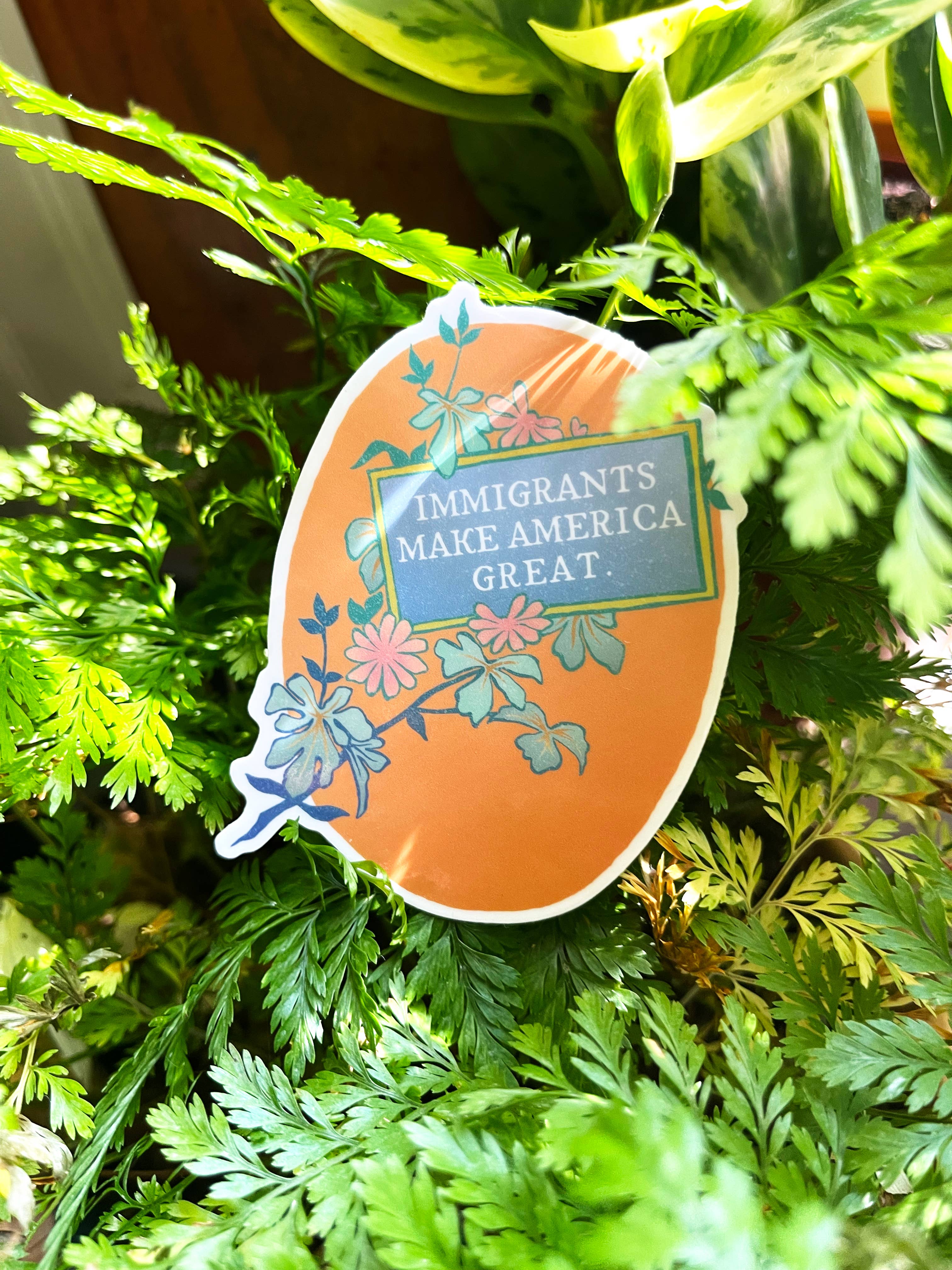 An oval-shaped sticker with an orange background featuring green leaves and the text 'Immigrants Make America Great'. The sticker has a adhesive back for placing on various surfaces.