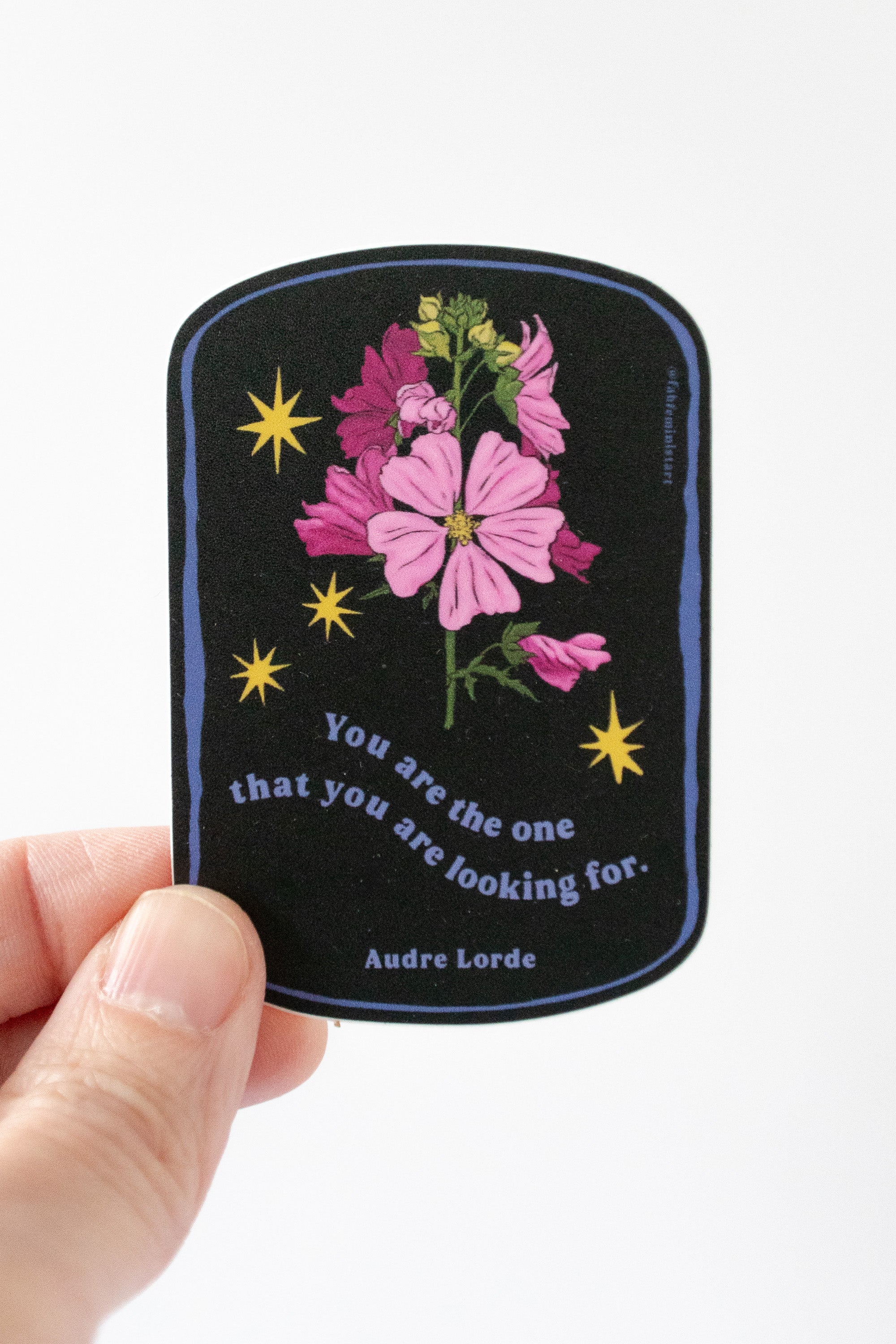 A hand holding a sticker with Audre Lorde's quote 'You are the one that you are looking for.' The sticker has a black background with pink flowers and yellow stars.